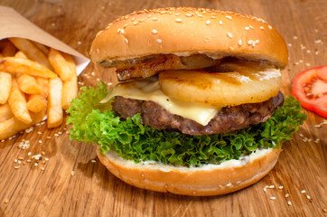 Big tasty burger on the wooden table with becon and french fries. Delicious dinner with cheese, tomatoos and salad. Fast food with beer.