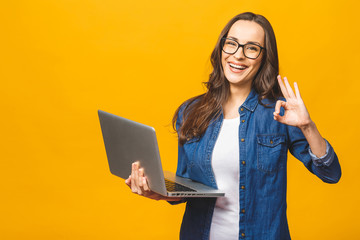 Portrait of a cheerful young girl holding laptop computer and showing ok isolated over yellow background.
