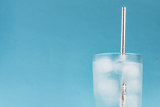 Close Up Of Pure Water With Ice And Silver Paper Straw In Tall Transparent Glass And Sun Glare On Blue  Background. Copy Space