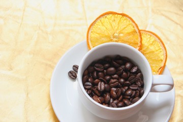 Cup and saucer, coffee beans, slices of dried orange on a gold background. A white cup and saucer with roasted coffee beans, with two pieces of dried orange and a couple of coffee beans on the saucer