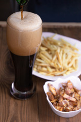 Vertical. The glass is filled with dark beer from the bottle, french fries and snacks in the background. Men's set to watch a football match or a party. Side view
