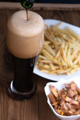 Vertical. The glass is filled with dark beer from the bottle, french fries and snacks in the background. Men's set to watch a football match or a party