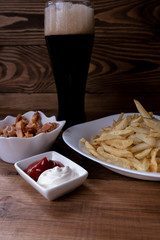 Vertical. Dark beer in a glass and snacks to it: french fries, pork ears and sauces on a dark wooden background. Men's set to watch a football match