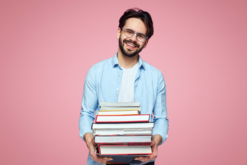 Young smiling hipster boy in blue shirt holds heap of books borrowed from library, isolated over pink backdrop