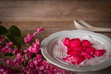Thapthim krop, mock pomegranate seeds in coconut and syrup in the white bowl on the wood table there are flower and spoon placed around.