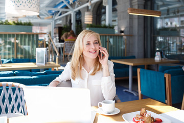 Happy young businesswoman or manager of large company talking on the phone with friends or colleagues discussing plans for the weekend sitting at table in cafe with laptop.