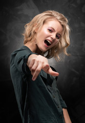 Beautiful young blonde girl with disheveled hair and nude makeup, wearing a shirt and jeans emotionally posing on a gray background. She screams and points a finger.