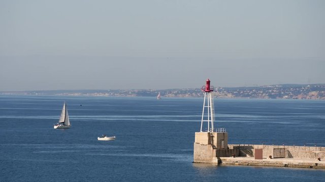 View from Marseille over the Mediterranean Sea. There is a sailboat passing by and a lighthouse at the end of a pier. Filmed in february.
