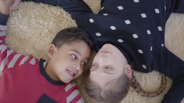 Portrait cute african american boy and blond caucasian girl lying on the floor on the beige fluffy carpet and looking at each other with love. Top view. Rest of happy children.