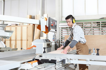 carpenter man work in the joinery, cut a wooden board with circular saw machine, protected with ear...