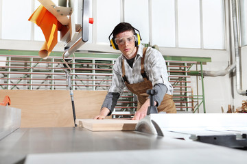 carpenter man work in the joinery, cut a wooden board with circular saw machine, protected with ear...