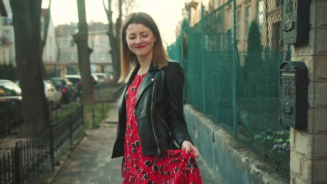 Nice Caucasian young woman in red long dress and black leather jacket walking through the street, dancing, laughing. Positive emotions. Outdoors. Happiness. Spring.