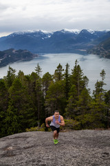 Naklejka na ściany i meble Fit and Muscular Young Man is Running up the Mountain during a cloudy day. Taken on Chief Mountain in Squamish, North of Vancouver, BC, Canada.