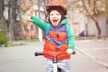 A small cool boy in a helmet and red vest shows thumb up and rides a running bike with a blue backpack on his back.