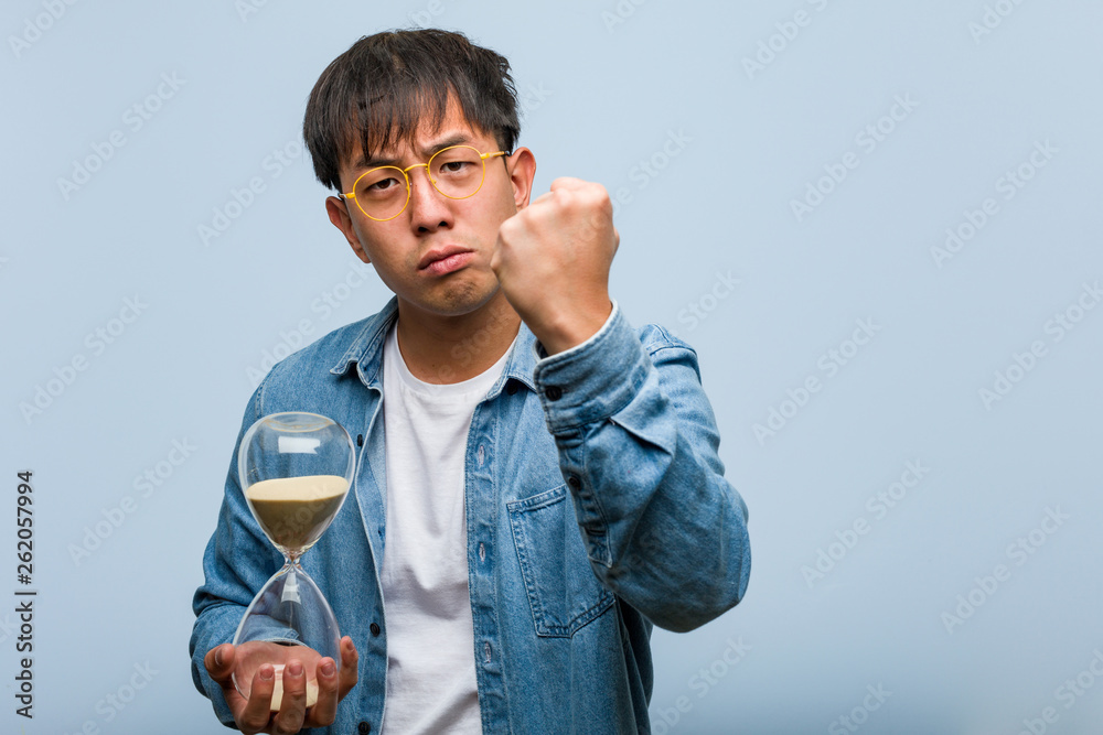 Wall mural young chinese man holding a sand timer showing fist to front, angry expression