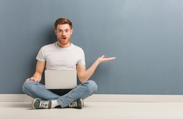 Young redhead student man sitting on the floor holding something on palm hand. He is holding a laptop.