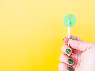 A woman with green painted nails with a green lollipop in her hand and yellow background