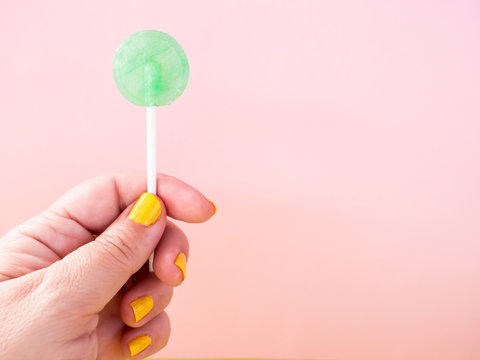 A woman with yellow painted nails with a green lollipop in her hand and pastel pink background