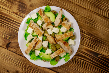 Tasty salad with fried chicken breast, green olives, feta cheese, avocado, lettuce and olive oil on wooden table. Top view