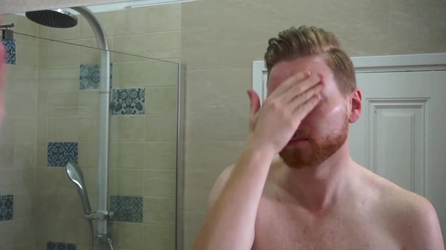 Young caucasian man applying face care face cream on his face and neck