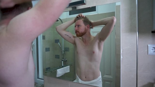 Young caucasian man fixing his hair and watching himself in the mirror in the bathroom