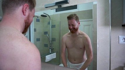 Young Skinny caucasian man trying to flex his muscles in front of the mirror in the bathroom wearing white towel