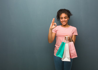 Young black woman cheerful and confident doing ok gesture. She is holding a shopping bags.