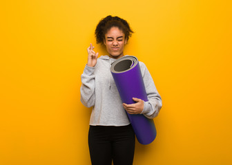 Young fitness black woman crossing fingers for having luck. Holding a mat.