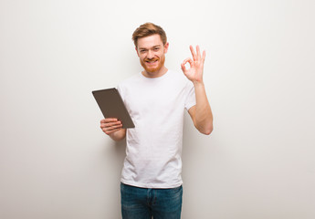 Young redhead man cheerful and confident doing ok gesture. Holding a tablet.