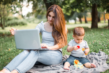 Mother with son sitting in the park and work on laptop.