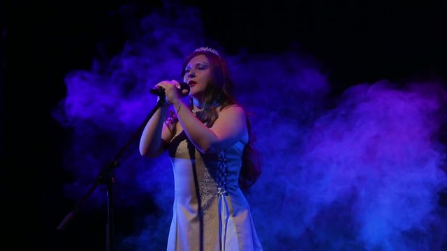 A young red-haired singer sings on stage with professional lighting and puffs of smoke