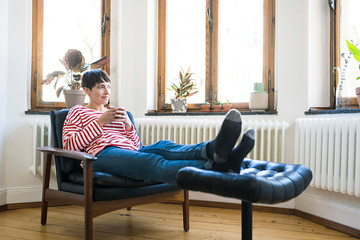 Short-haired woman relaxing in lounge chair holding coffee mug in stylish apartment