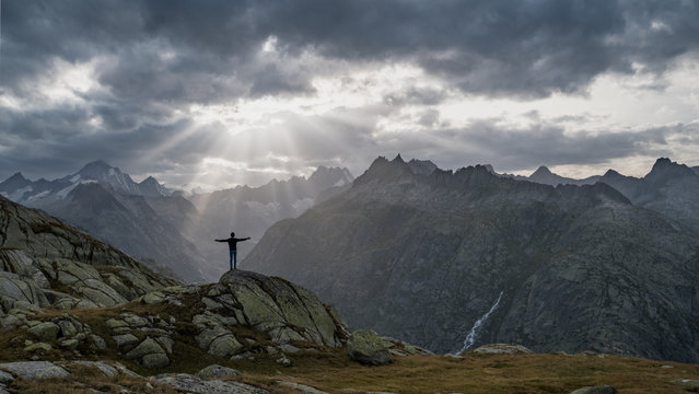 A Boy On A Trekking Trip Enjoys An Incredible View Of Sun Rays Breaking Through Thick Dark Clouds Over A Valley, Posing In 