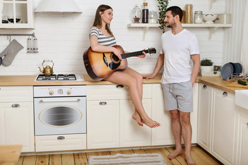 Smiling young beautiful woman singing and playing on acoustic guitar to her boyfriend or husband sitting in kitchen of new home. Happy family together concept