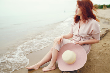 Fototapeta na wymiar A young redheaded girl in a large round hat and pink dress sitting on the beach near ocean