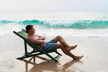 Young man sitting and relaxing on sun lounger in water at the beach in the sunshine. Blue sea waves at background