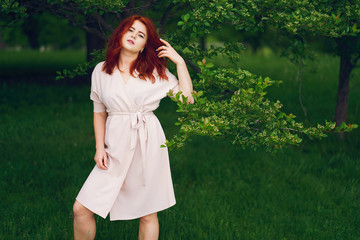 A young redheaded girl in a pink dress standing in a summer green park near trees