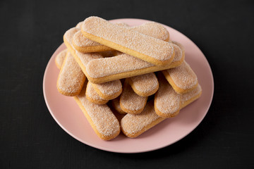 Savoiardi or ladyfingers cookies on pink plate over black background, side view.