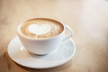 White cappuccino cup with with latte art heart on light brown wood background lit by bright morning sunlight