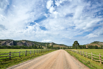 Rustic summer landscape. The road to the field. Cloudy sky. Mountains and hills in the background. Fence along the road. - 262091340