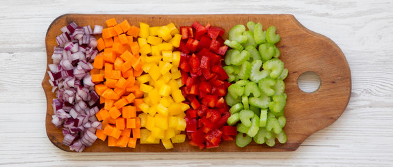 Chopped fresh vegetables (carrot, celery, red onion, colored peppers) arranged on cutting board on a white wooden surface, top view. Flat lay, from above, overhead. Copy space.