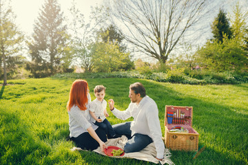 beautiful and stylish redhead mom in a white blouse sits on the grass with her beautiful man dressed in a white shirt and blue jeans; they play with their little sweet son who eats grapes