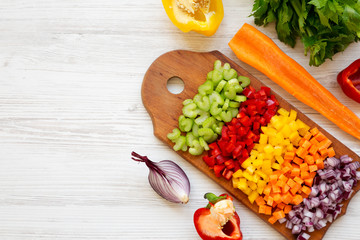 Chopped fresh vegetables (carrot, celery, onion, colored peppers) arranged on cutting board on a white wooden background, top view. Copy space.