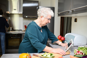 happy senior active woman cooking at home in modern kitchen and looking for recipe on a internet computer tablet