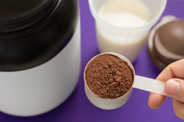 Whey Protein. Point of view of hand holding measuring scoop. Chocolate flavour. Color background: violet.