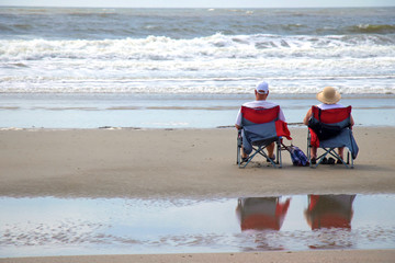 An unidentifiable mature man and woman sit with backs to the camera in colorful beach chairs on the sand facing the surf in soft morning or afternoon light.