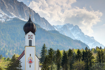 Close-up of small Bavarian domed village church with white facade and clock tower with snowy Zugspitze peak and heap clouds in background in spring sunshine, Wetterstein Grainau Bayern Germany Europe