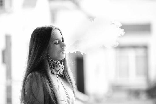 Vape teenager. Young pretty white girl in casual clothing smoking an electronic cigarette on the street in the spring. Bad habit. Vaping activity. Close up. Black and white.