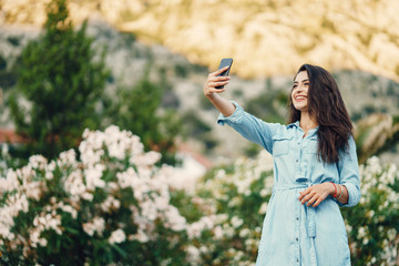 A beautiful young girl in a blue dress standing near the flower tree and use the phone