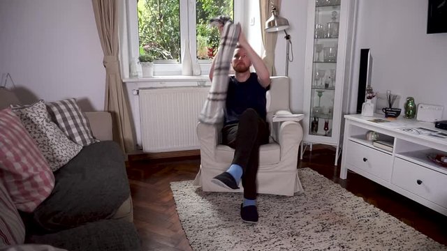 Intelligent young man puts a warm blanket on his legs reading a book in cozy armchair while spending pastime in loft apartment